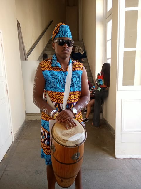 Participante com roupa de estampa afro tocando um tambor